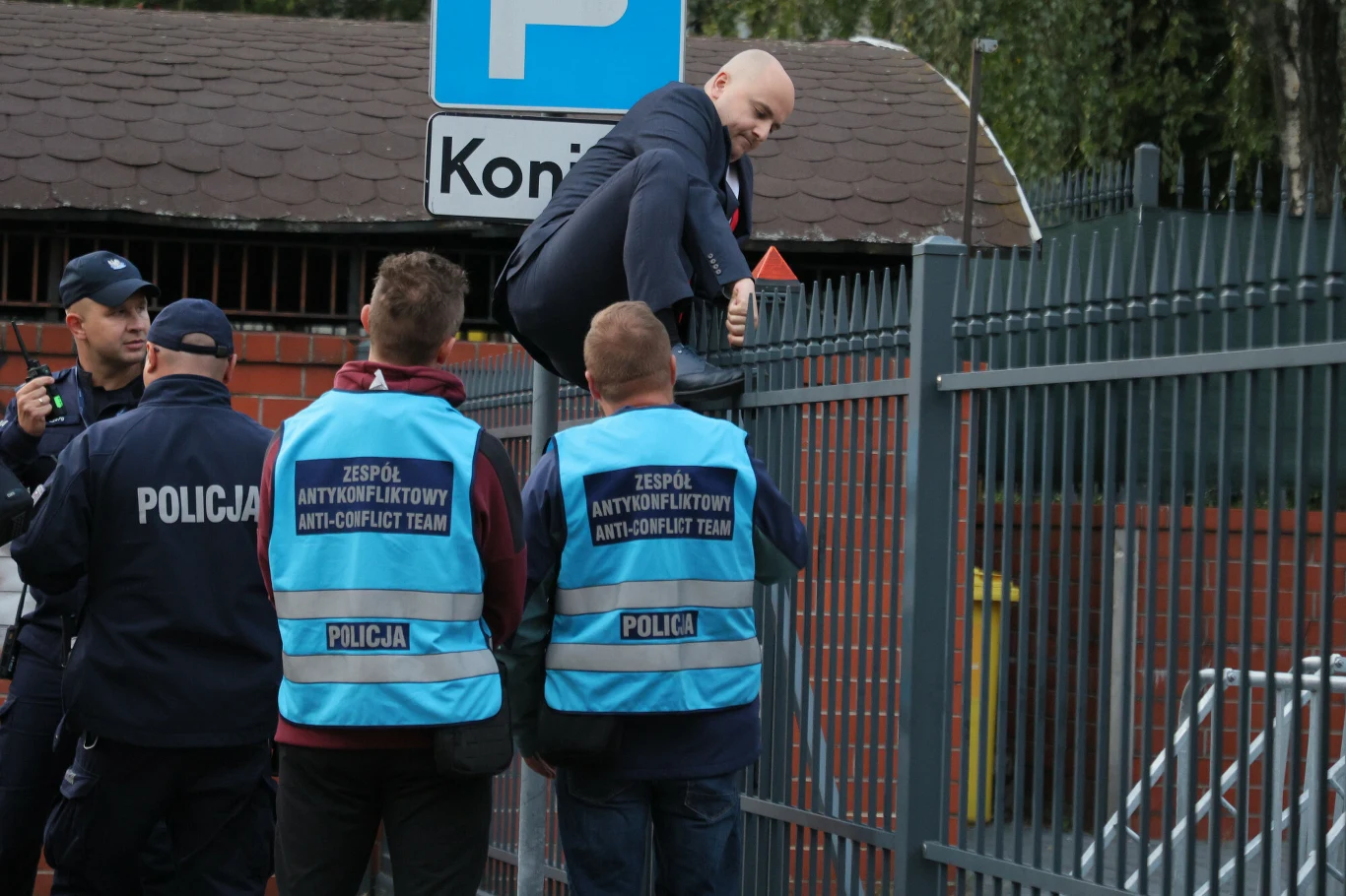 Funkcjonariusze policji, w tym członkowie zespołu antykonfliktowego, zatrzymują mężczyznę w garniturze, który wspina się przez metalowe ogrodzenie.