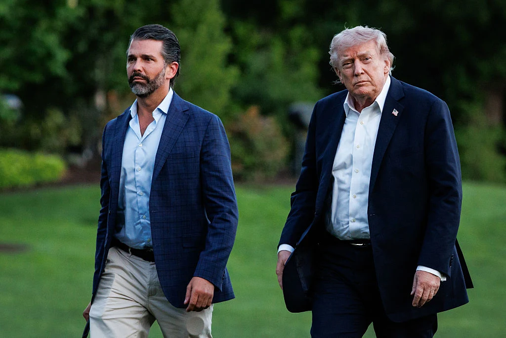 Donald Trump Jr. i jego ojciec, prezydent USA Donald Trump Donald Trump i Donald Trump Jr. idą w ciemnych marynarkach po zielonym trawniku, wokół drzewa i krzewy.
