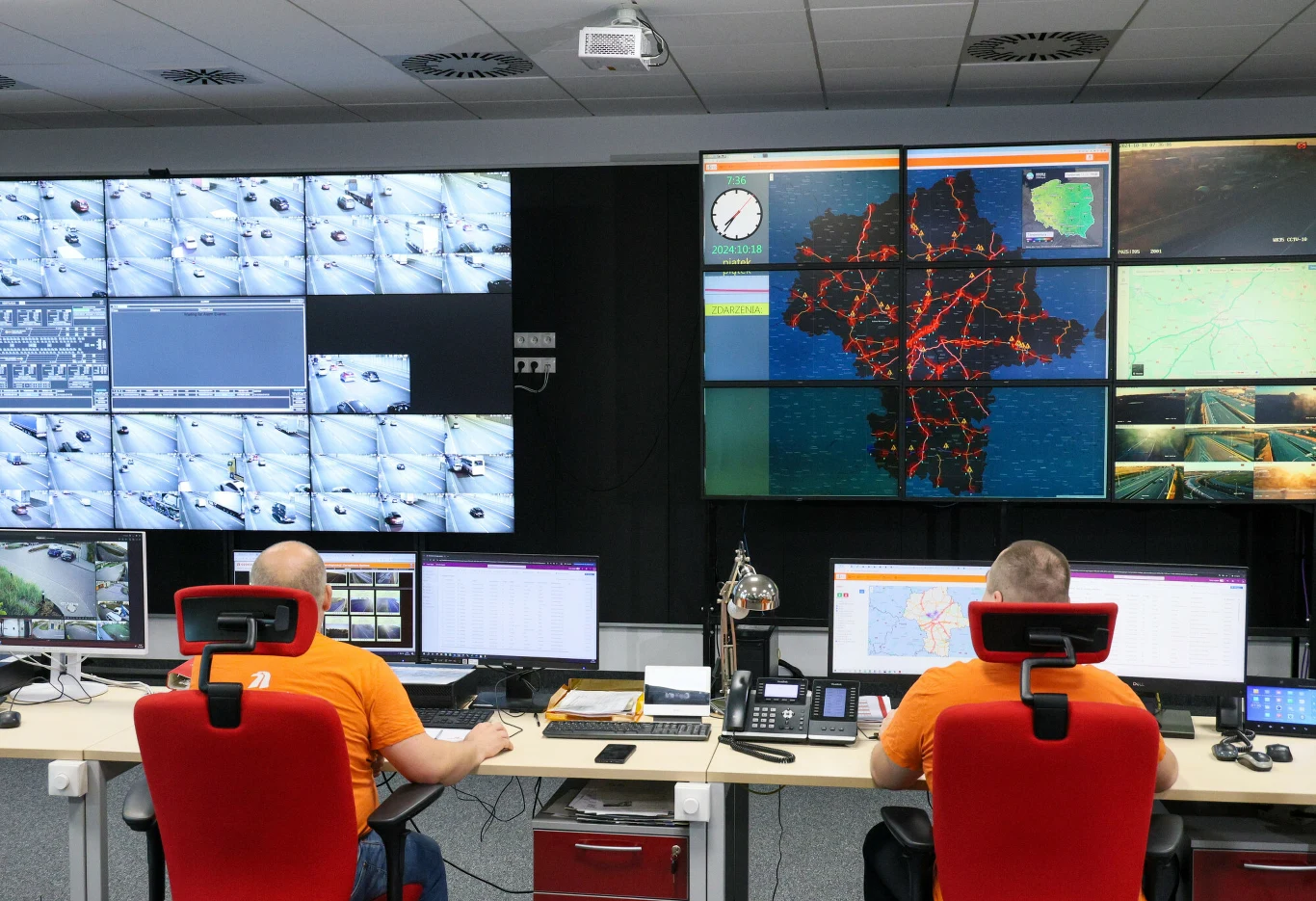 Centrum Zarzadzania Tunelem POW Warszawa. Tunel na S2 to najdłuższa taka przeprawa w Polsce Dwóch operatorów w pomarańczowych koszulkach pracuje przy dużych stanowiskach przed ścianą monitorów, na których wyświetlane są obrazy z kamer przemysłowych oraz wizualizacje terenu i mapy z zaznaczonymi ścieżkami ruchu.