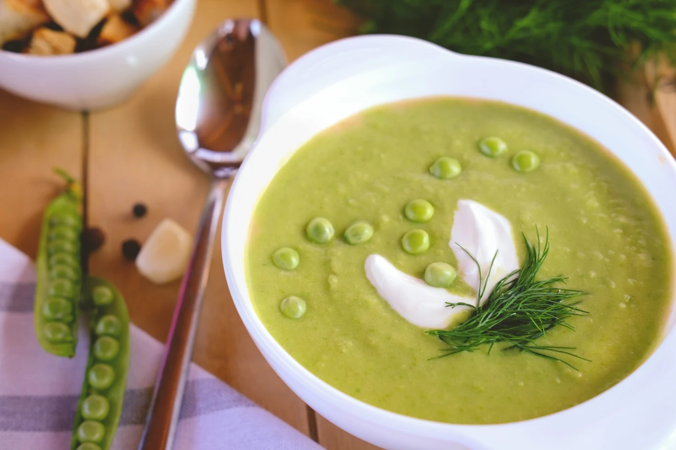 Zupa krem z zielonego groszku to wybawienie w szalone dni i ulubiony przepis zabieganych. 25 minut i masz obiad jak z restauracji Kremowa zupa z zielonego groszku z kleksem śmietany, koperkiem i groszkiem, podana w białym talerzu.