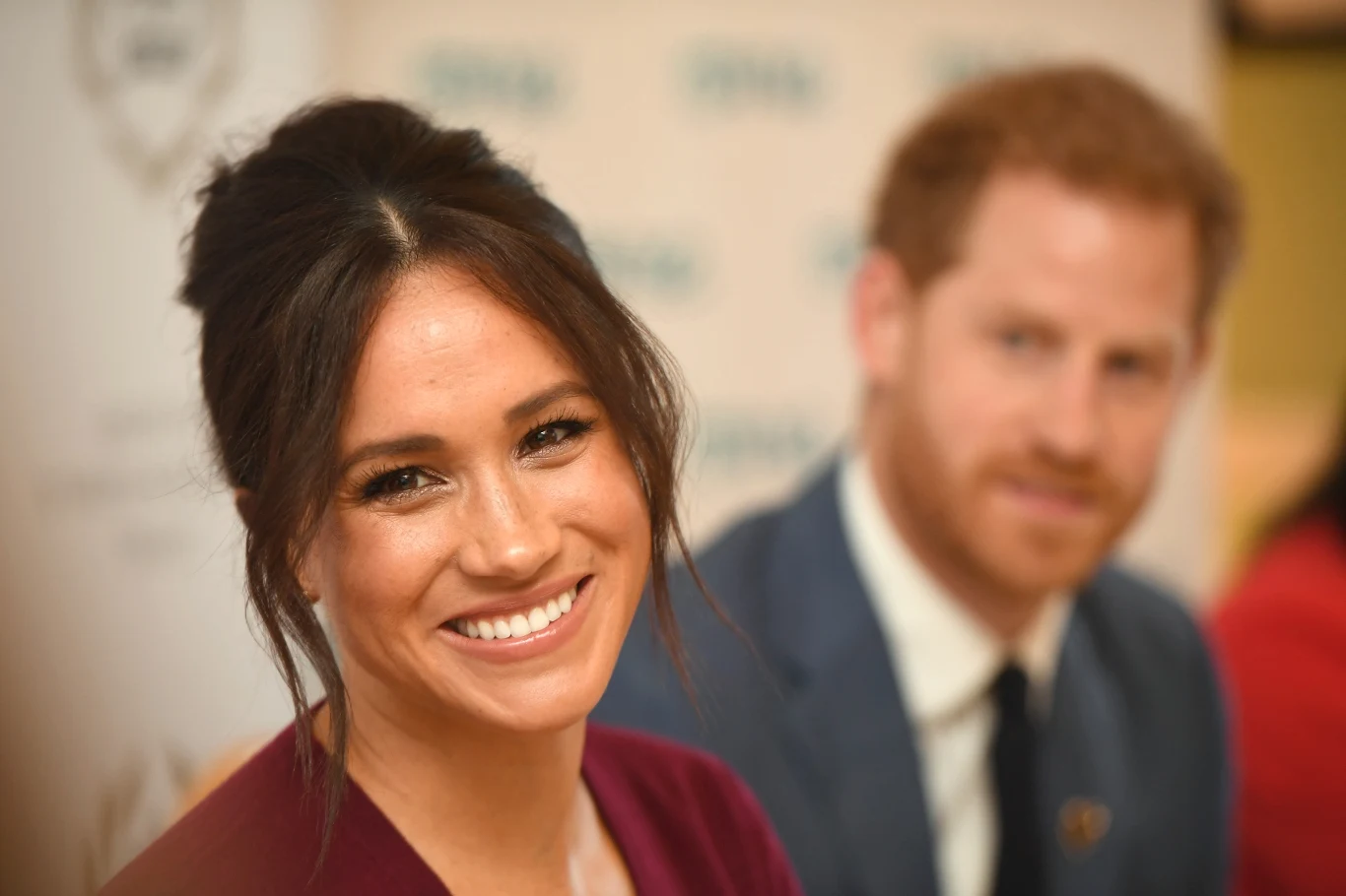 Ochrona ma także analizować sytuację Meghan Markle Uśmiechnięta kobieta z ciemnymi włosami spiętymi w luźny kok siedzi przy stole, w tle mężczyzna w garniturze i krawacie, tło rozmyte z widocznym logo na białym tle.