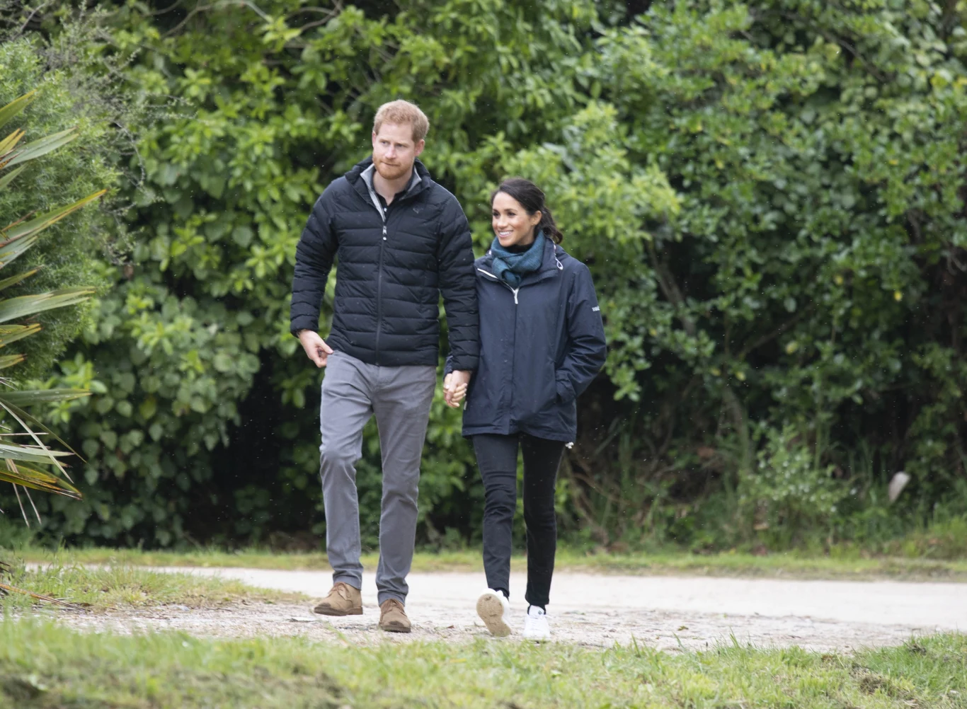 Osoby śledzące sytuację Meghan i Harry'ego zastanawiają się, jak dalej potoczą się ich losy Para idzie razem alejką wśród zielonej roślinności, trzymając się za ręce i uśmiechając się do siebie.