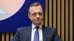 Mężczyzna w ciemnym garniturze i niebieskim krawacie siedzi na fotelu, z wyrazem skupienia na twarzy, trzymając czarny notes na kolanach; za nim tło z pionowymi liniami i wycinkiem dużego ekranu.
