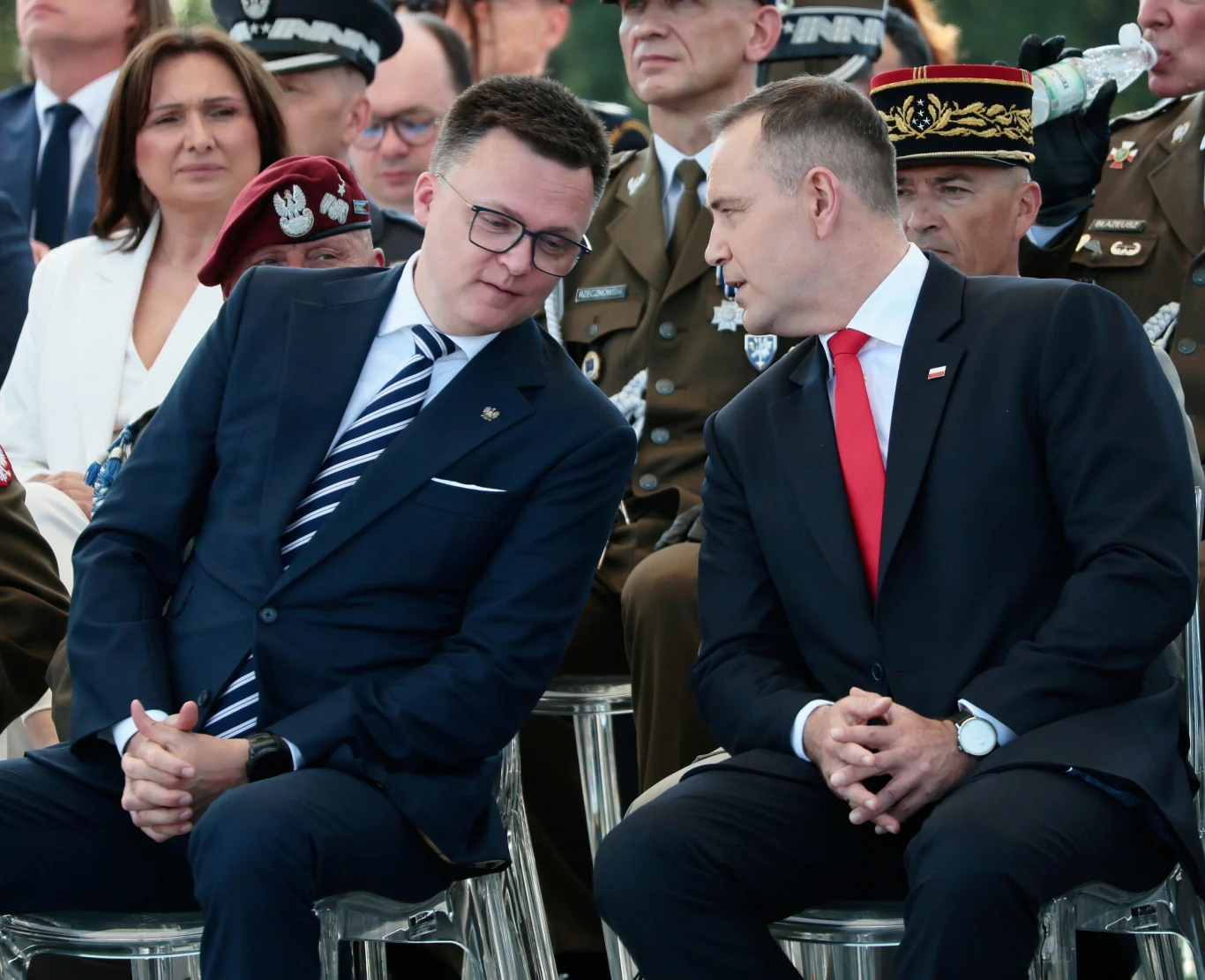 Dwóch elegancko ubranych mężczyzn w garniturach rozmawia ze sobą, siedząc w pierwszym rzędzie obok wojskowych w mundurach podczas oficjalnego wydarzenia.