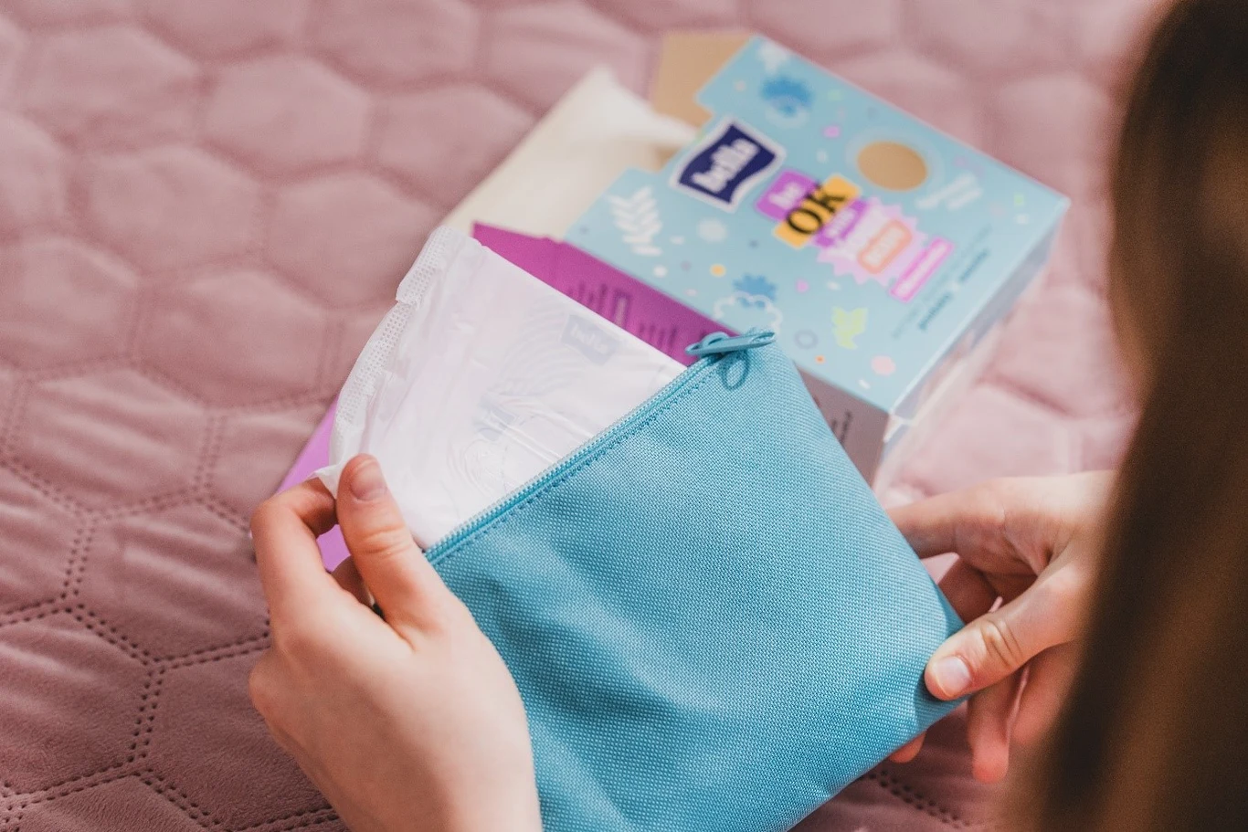 W przypadku Bella for Teens można skorzystać z gotowych zestawów miesiączkowych, takich jak OKresówka Bella for teens, stanowiących starannie skomponowany niezbędnik Ręce wkładają podpaskę higieniczną do niebieskiej kosmetyczki, obok znajduje się otwarte opakowanie podpasek leżące na różowej pikowanej powierzchni.