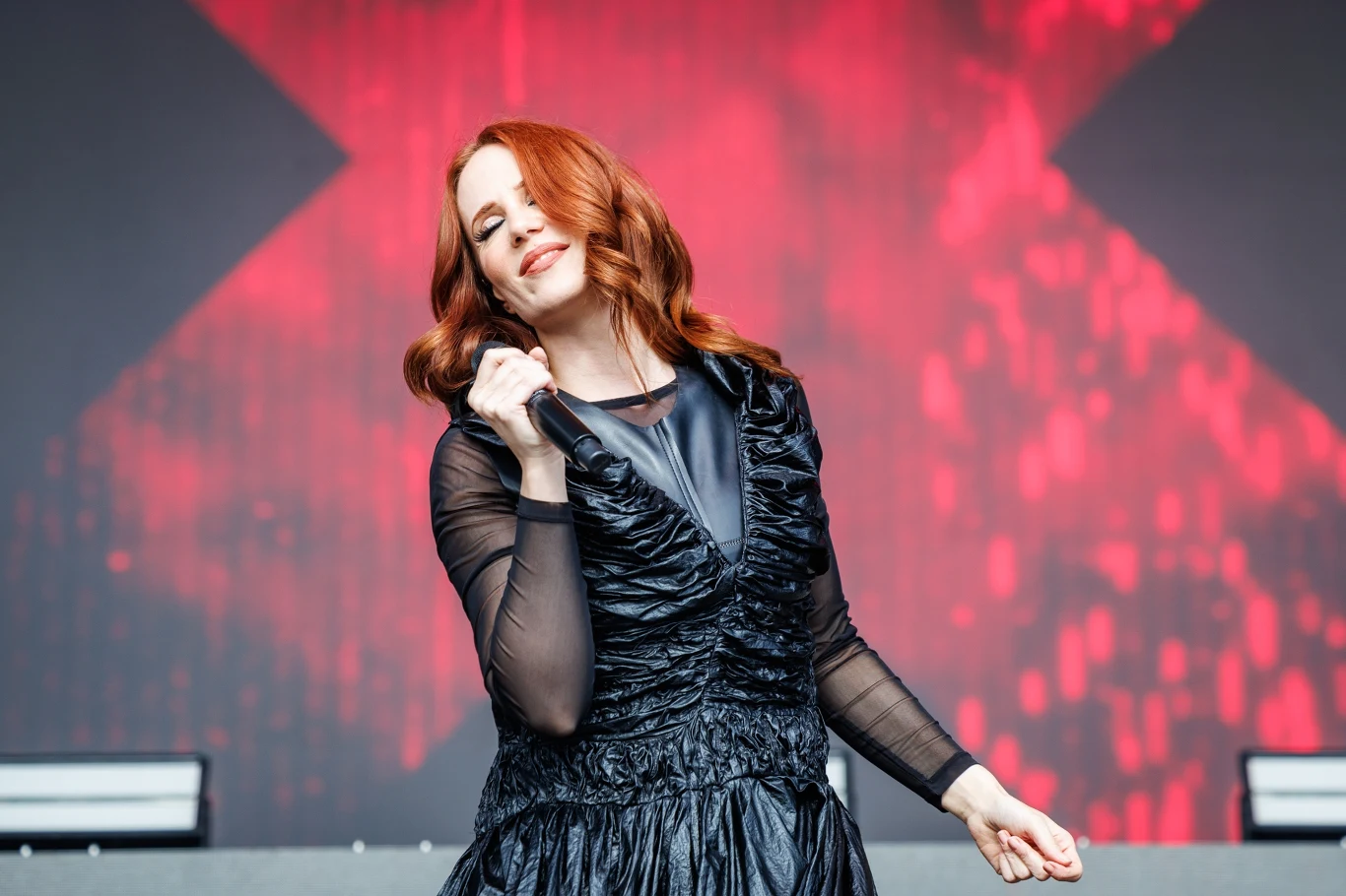 Simone Simons (Epica) w akcji Simone Simons z Epica śpiewa w czarnej, błyszczącej sukni na tle sceny z czerwono-czarną grafiką.