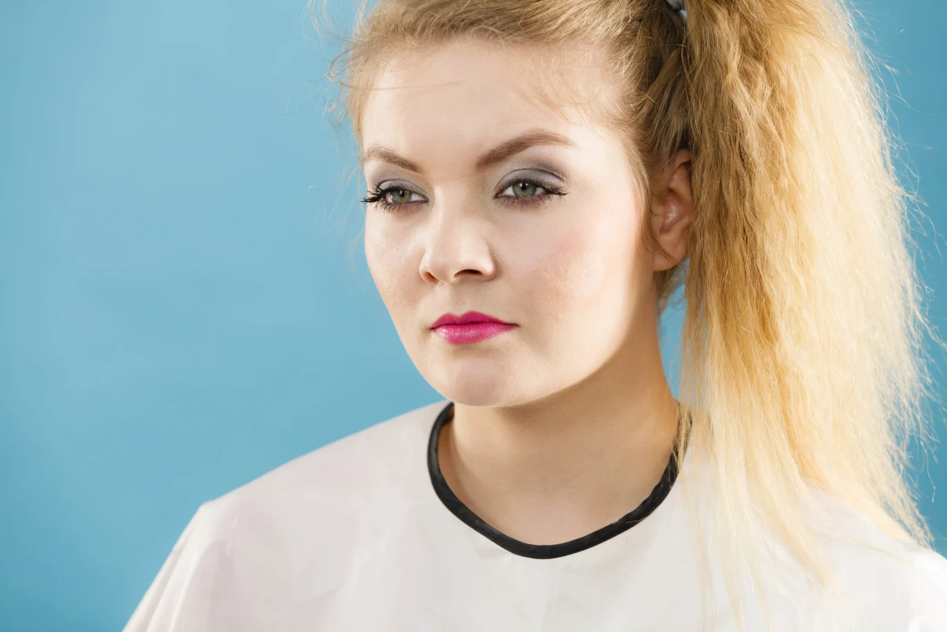 Karbowane włosy na jesień 2026. Trend lat 90. znowu modny Młoda kobieta z blond karbowanymi włosami w wysokim kucyku, w białym fartuchu na tle błękitnej ściany.
