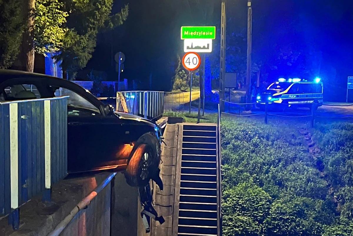 Czarny samochód osobowy zawieszony nad krawędzią betonowej skarpy, z widocznym uszkodzeniem przedniego koła i błotnika, w tle radiowóz policyjny z włączonymi światłami oraz znak drogowy z nazwą miejscowości Międzylesie i ograniczeniem prędkości do 40 k...