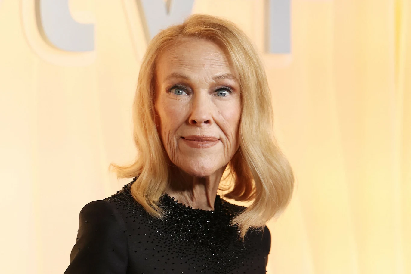 Catherine O'Hara Catherine O'Hara o blond włosach, ubrana w czarną sukienkę z ozdobnym kołnierzem, pozuje na tle jasnej, żółtej ściany z napisami.
