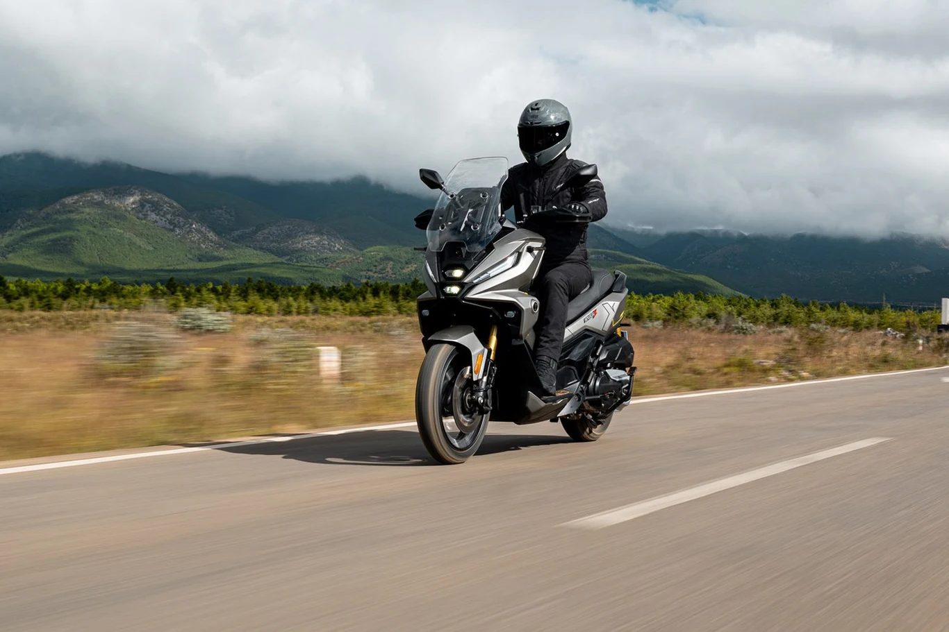 Dwucylindrowy silnik o pojemności 398 cm³ rozwija 42 KM i współpracuje z automatyczną przekładnią CVT. Motocyklista w czarnym ubraniu i kasku jedzie nowoczesnym motocyklem po pustej asfaltowej drodze, w tle widoczne zielone wzgórza oraz niskie, przesuwające się chmury.