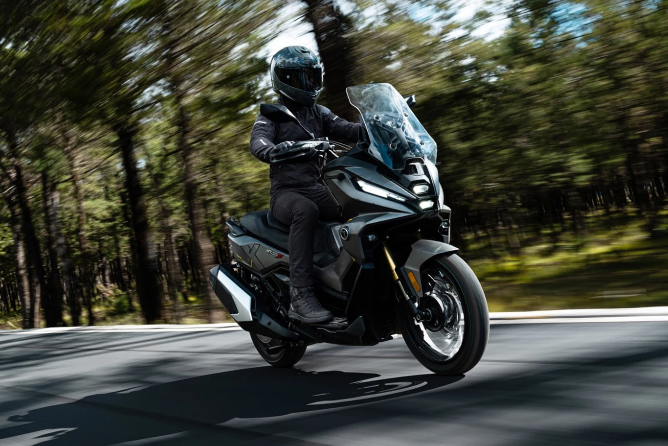 Wyposażenie obejmuje m.in. elektrycznie regulowaną szybę, podgrzewane manetki i kanapę oraz 7-calowy ekran TFT. Motocyklista w czarnym stroju i kasku jedzie nowoczesnym skuterem po asfaltowej drodze otoczonej lasem.