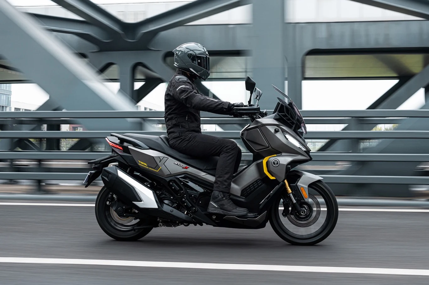 Voge SR450X łączy wygodę miejskiego skutera z rozwiązaniami zaczerpniętymi z motocykli turystycznych. Motocyklista w czarnym kombinezonie i kasku prowadzi nowoczesny skuter, przemieszczając się po miejskim moście z metalową konstrukcją w tle.
