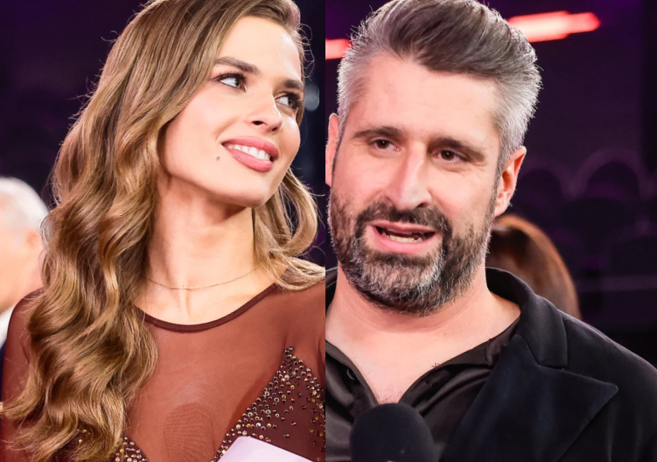 Dwie osoby w eleganckich strojach podczas wydarzenia, kobieta z długimi, falowanymi włosami i uśmiechem, mężczyzna z siwiejącymi włosami i brodą mówi do mikrofonu.