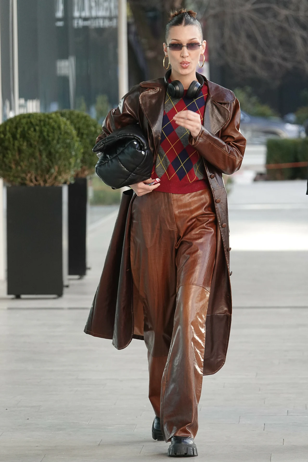 Bella Hadid w sweterku w szkocką kratę Kobieta ubrana w długi brązowy płaszcz z imitacji skóry, szerokie spodnie w tym samym kolorze, sweter w romby w odcieniach bordo i granatu, trzyma czarną torbę pod pachą, ma na szyi słuchawki i nosi okulary przeciwsłoneczne.