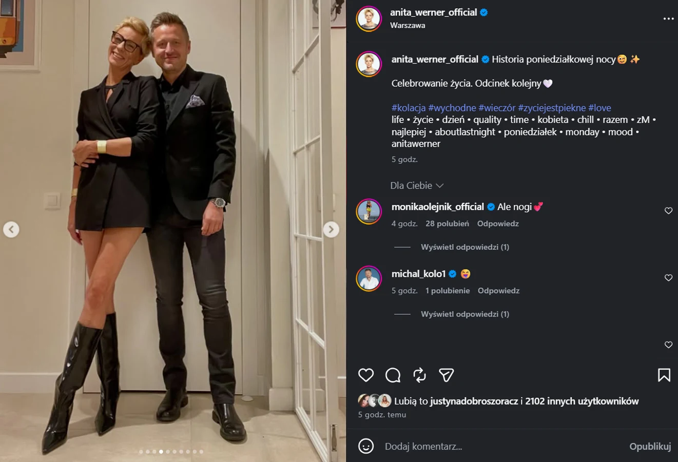 Uśmiechnięta para ubrana w eleganckie stylizacje, kobieta w czarnym, krótkim żakiecie i wysokich kozakach, mężczyzna w garniturze, stoją objęci na jasnym korytarzu.