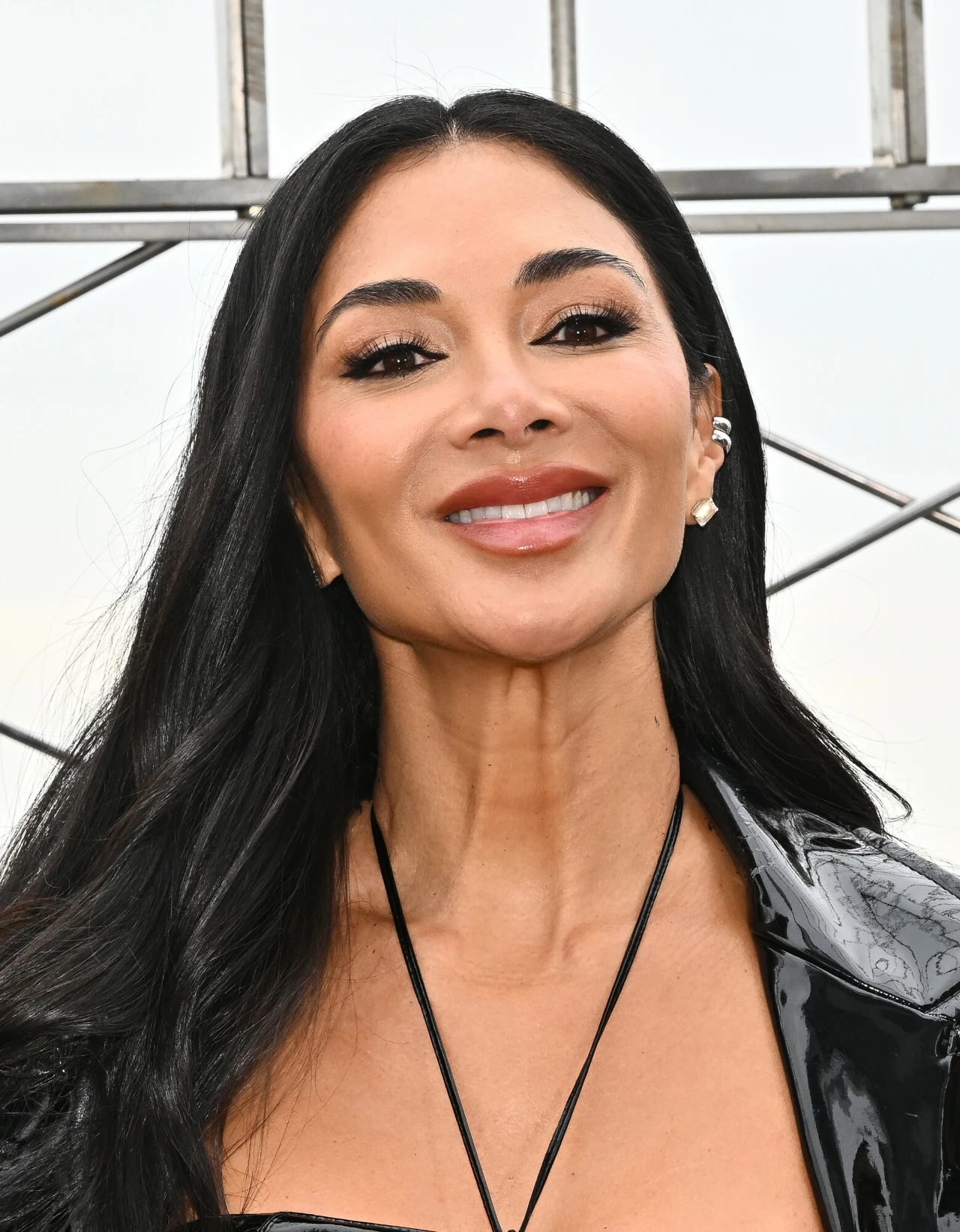 Nicole Scherzinger Uśmiechnięta kobieta z długimi, czarnymi włosami i wyrazistym makijażem, ubrana w błyszczącą czarną kurtkę i ozdobna kolczyk w uchu.