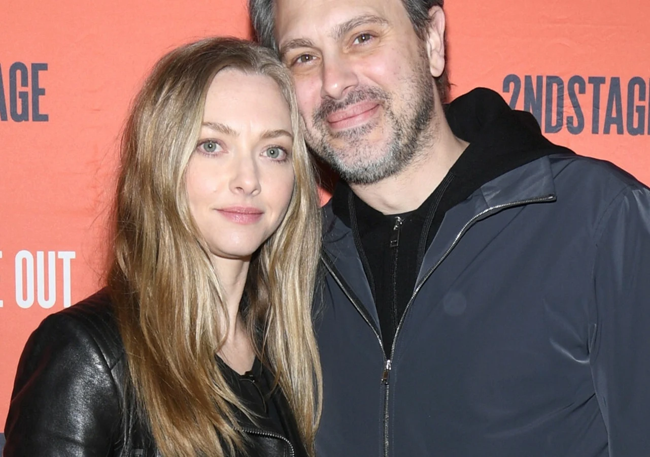 Amanda Seyfried i Thomas Sadoski Dwie osoby stoją obok siebie na tle pomarańczowej ścianki z logo, patrząc prosto w obiektyw; kobieta o jasnych włosach w czarnej kurtce oraz mężczyzna z zarostem w ciemnej kurtce z delikatnym uśmiechem.