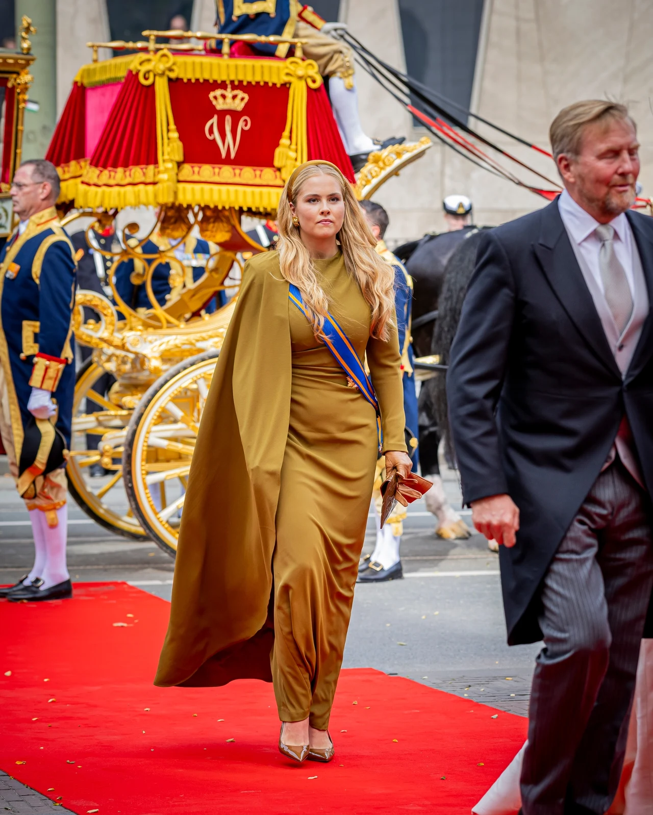 Catharina-Amalia jest następczynią holenderskiego tronu Kobieta w długiej oliwkowej sukni z peleryną i szarfą, idąca po czerwonym dywanie w otoczeniu elegancko ubranych osób oraz bogato zdobionego, złotego powozu z herbem.
