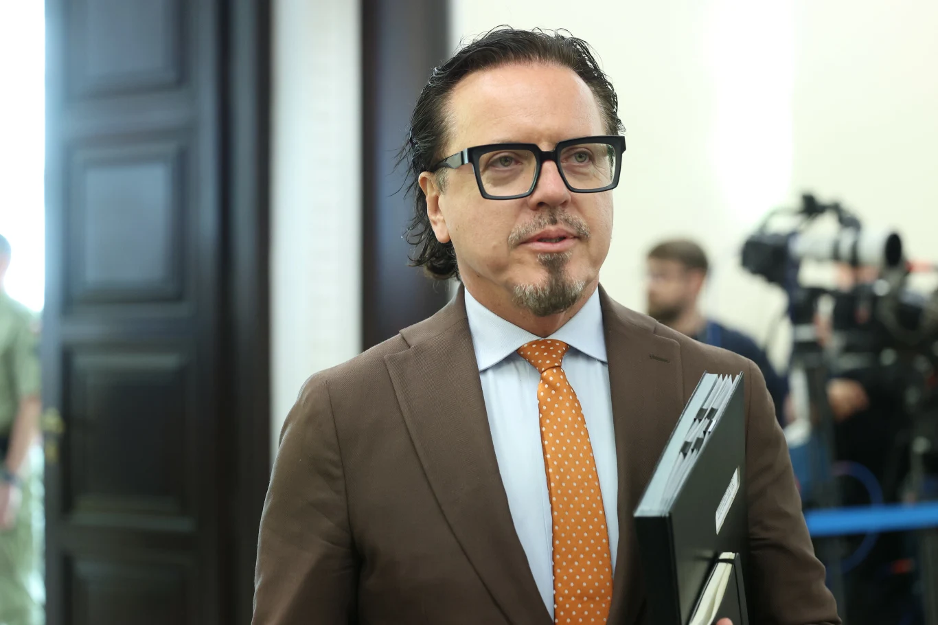 Minister aktywów państwowych Wojciech Balczun Wojciech Balczun w brązowym garniturze z pomarańczowym krawatem, trzyma czarny segregator na tle sprzętu filmowego.