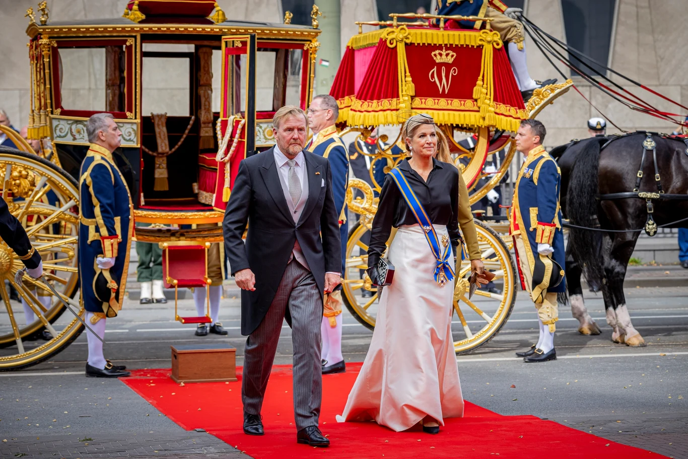Król i królowa Holandii zdecydowanie wyróżniali się stonowanymi kreacjami na tle barwnych stylizacji córek Para w eleganckich strojach maszeruje po czerwonym dywanie obok zdobionej, królewskiej karety w otoczeniu mundurowych służb ceremonialnych.