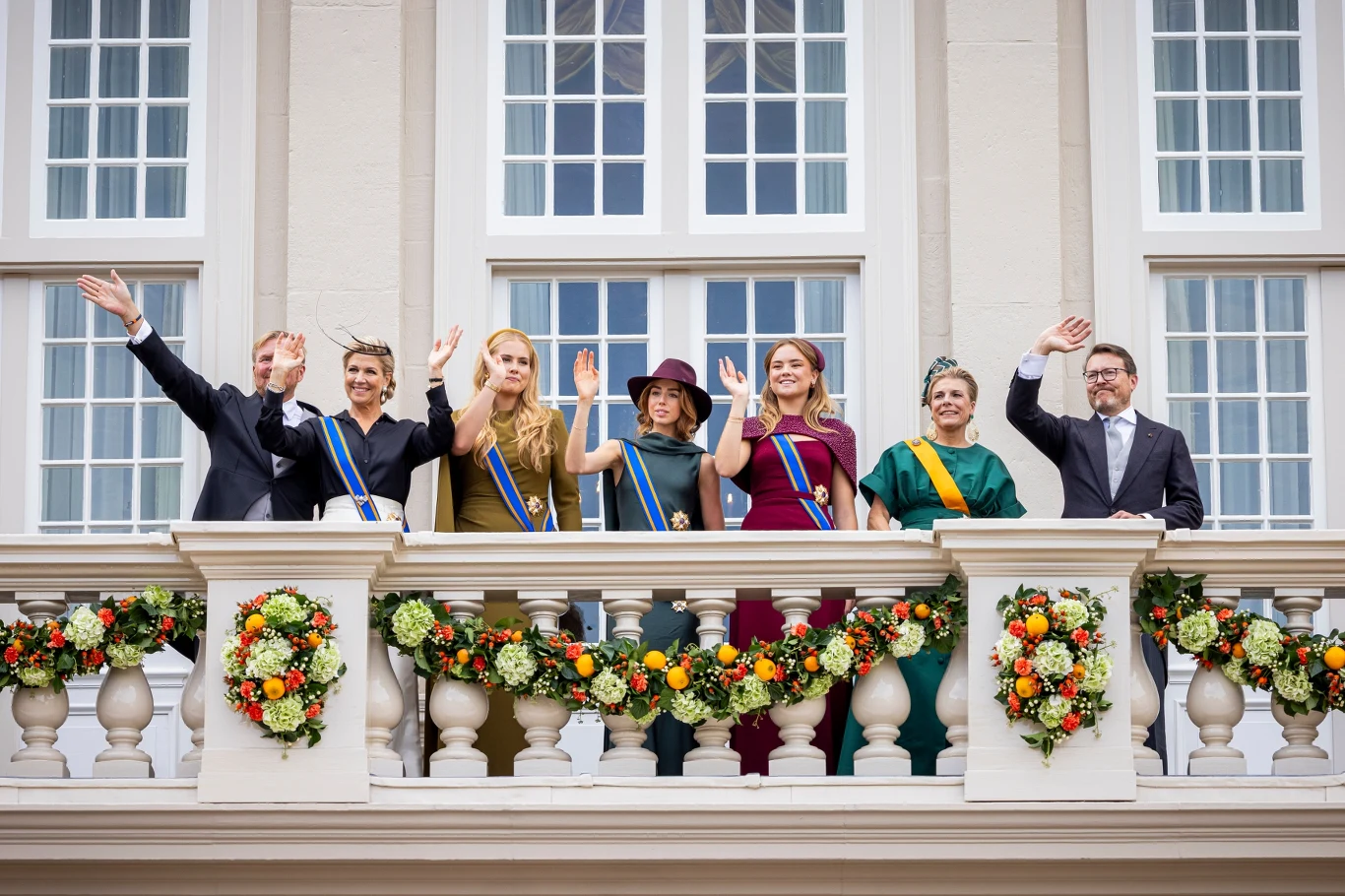 Królewska scena balkonowa podczas państwowych uroczystości jest tym momentem, na który zawsze czekają fani i poddani Grupa siedmiu elegancko ubranych osób stoi na udekorowanym balkonie, machając rękami w stronę zgromadzonych poniżej, w tle duże okna budynku i kompozycje kwiatowe zawieszone na balustradzie.