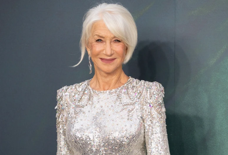 Helen Mirren Helen Mirren w srebrnej sukni z ozdobnymi ramionami, z siwymi włosami na tle ciemnej ściany.