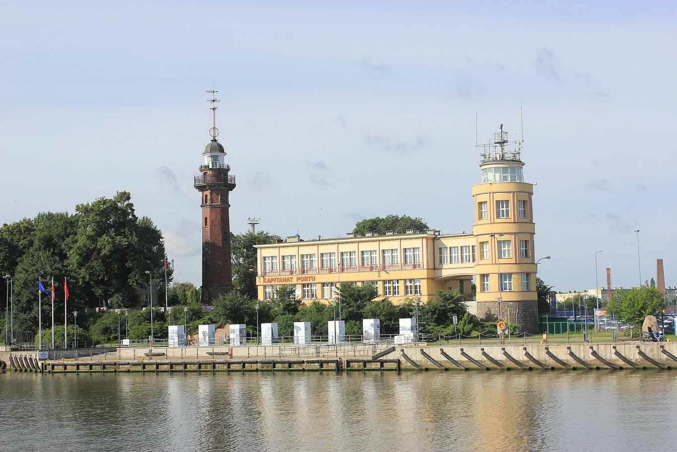 Koniec słynnej latarni w Gdańsku. Zostały ostatnie dni (Na zdjęciu: latarnia morska i kapitanat portu w Nowym Porcie) Latarnia morska i żółty budynek kapitanatu portu w Nowym Porcie nad wodą, otoczone zielenią i masztami z flagami.