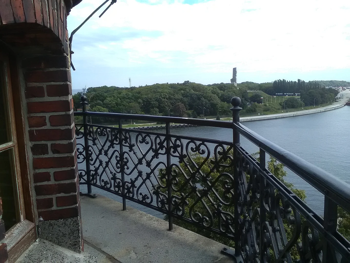 Widok z zabytkowej latarni w Nowym Porcie na Westerplatte Żelazna balustrada balkonu przy ceglanej ścianie budynku, z widokiem na rzekę i zieloną skarpę, w oddali widoczna wysoka wieża na tle nieba.