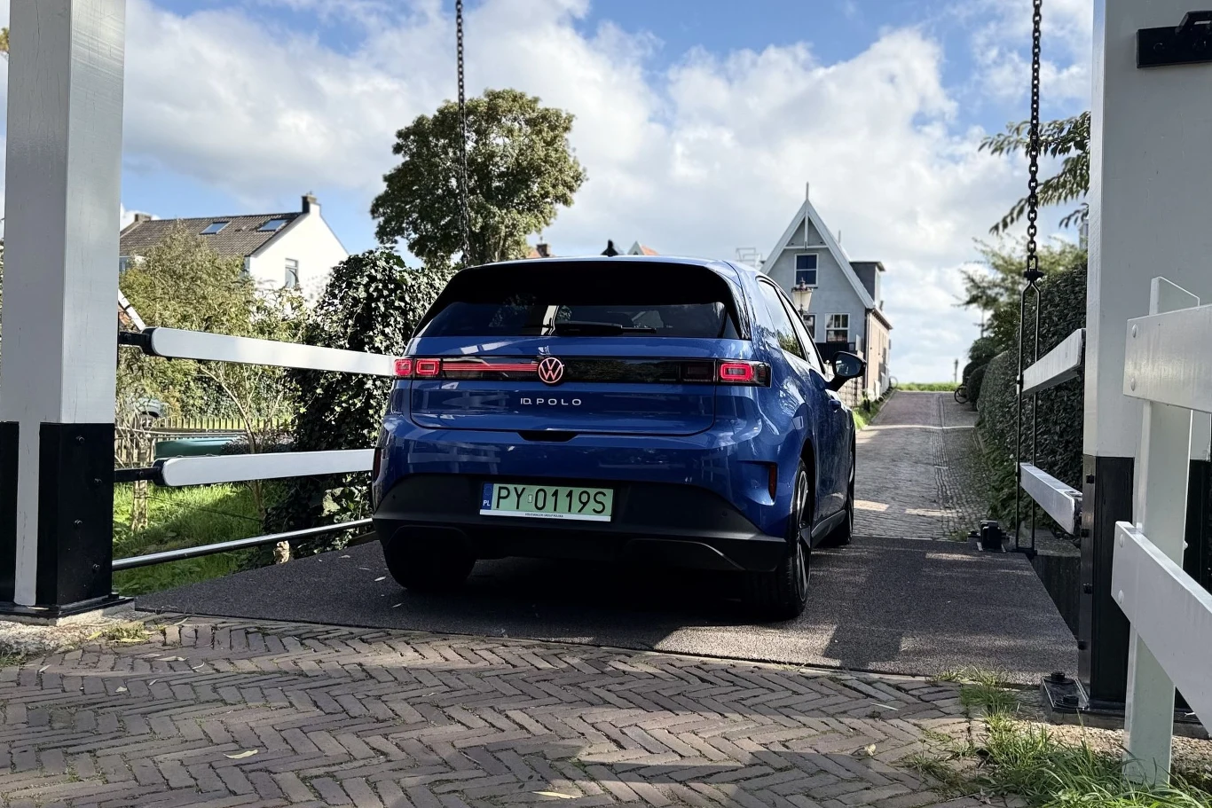 Niebieski Volkswagen ID.3 z zieloną tablicą rejestracyjną poruszający się przez most na tle białego domu z charakterystycznym, wysokim dachem.