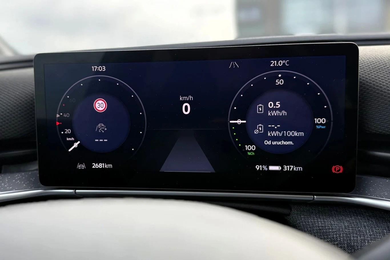 Cyfrowy kokpit samochodu z wyświetlonym limitem prędkości 30 km/h, aktualną prędkością zero, poziomem naładowania baterii 91% oraz zasięgiem 317 km, przy temperaturze zewnętrznej 21°C.