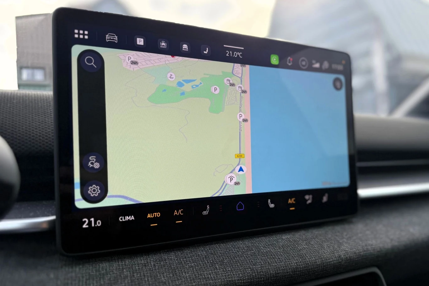 Ekran systemu multimedialnego samochodu z włączoną nawigacją, prezentujący mapę z drogą biegnącą wzdłuż linii brzegowej, przy której oznaczono kilka punktów parkingowych.