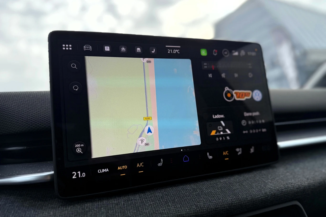 Duży ekran systemu multimedialnego w samochodzie z wyświetloną nawigacją GPS i panelem sterowania klimatyzacją, otoczony nowoczesnym wnętrzem pojazdu.