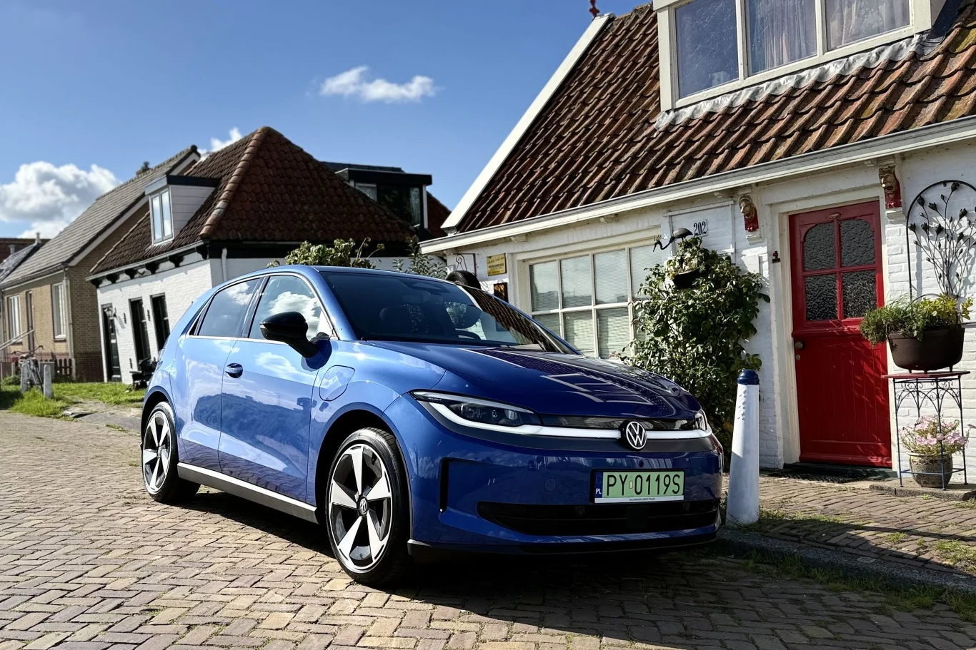 Niebieski Volkswagen Golf zaparkowany na brukowanej ulicy przy białym domu z czerwonymi drzwiami i kwiatami w donicach.