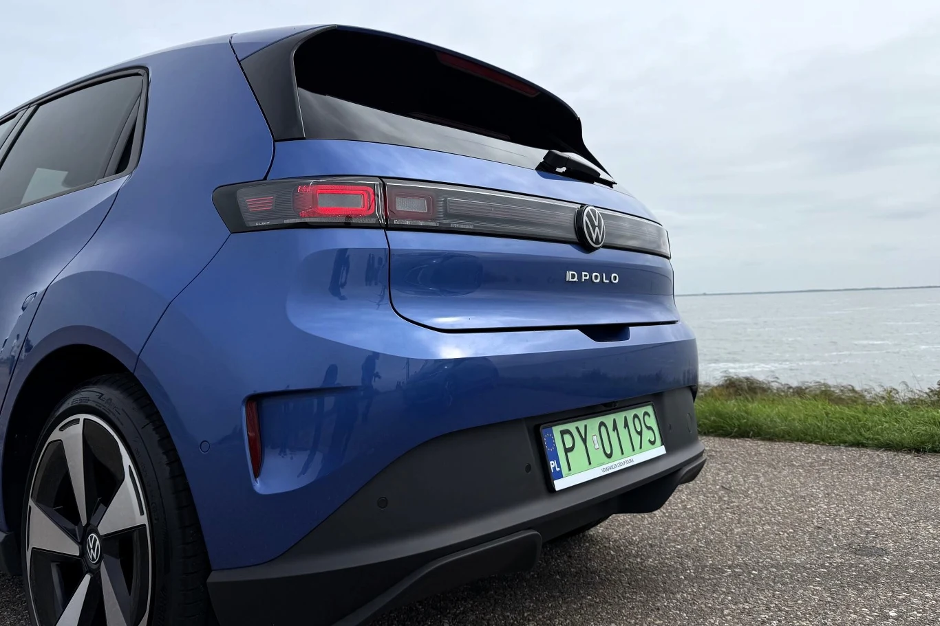 Błękitny samochód Volkswagen Polo zaparkowany przy drodze nad brzegiem wody, widoczny tył pojazdu z nowoczesnym, poziomym oświetleniem LED i zieloną tablicą rejestracyjną.