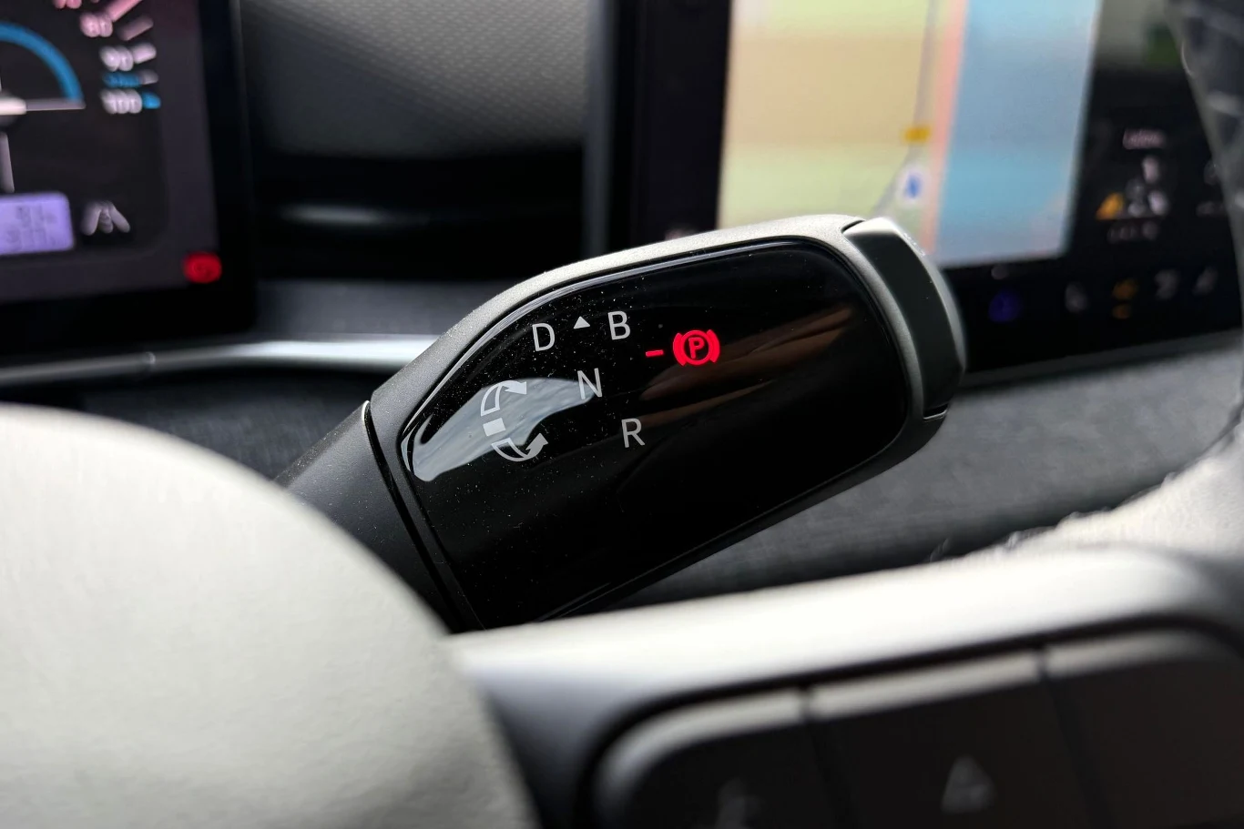 Dźwignia zmiany biegów w samochodzie elektrycznym z widocznymi oznaczeniami trybów jazdy oraz ikona zaciągniętego hamulca postojowego, w tle fragmenty wyświetlaczy elektronicznych.