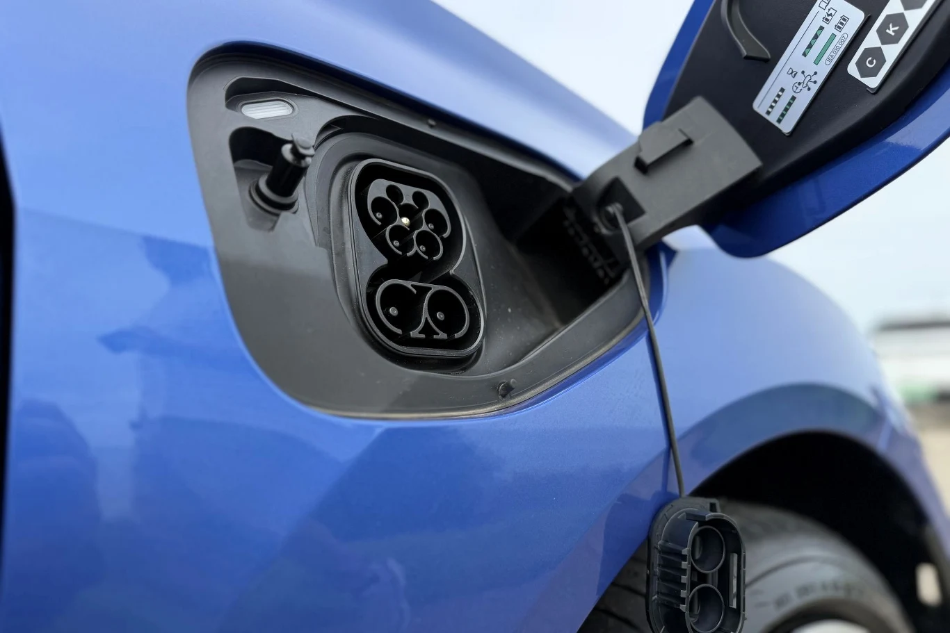 Gniazdo ładowania samochodu elektrycznego w niebieskim nadwoziu z otwartą klapką portu, widocznymi stykami i etykietą informacyjną.