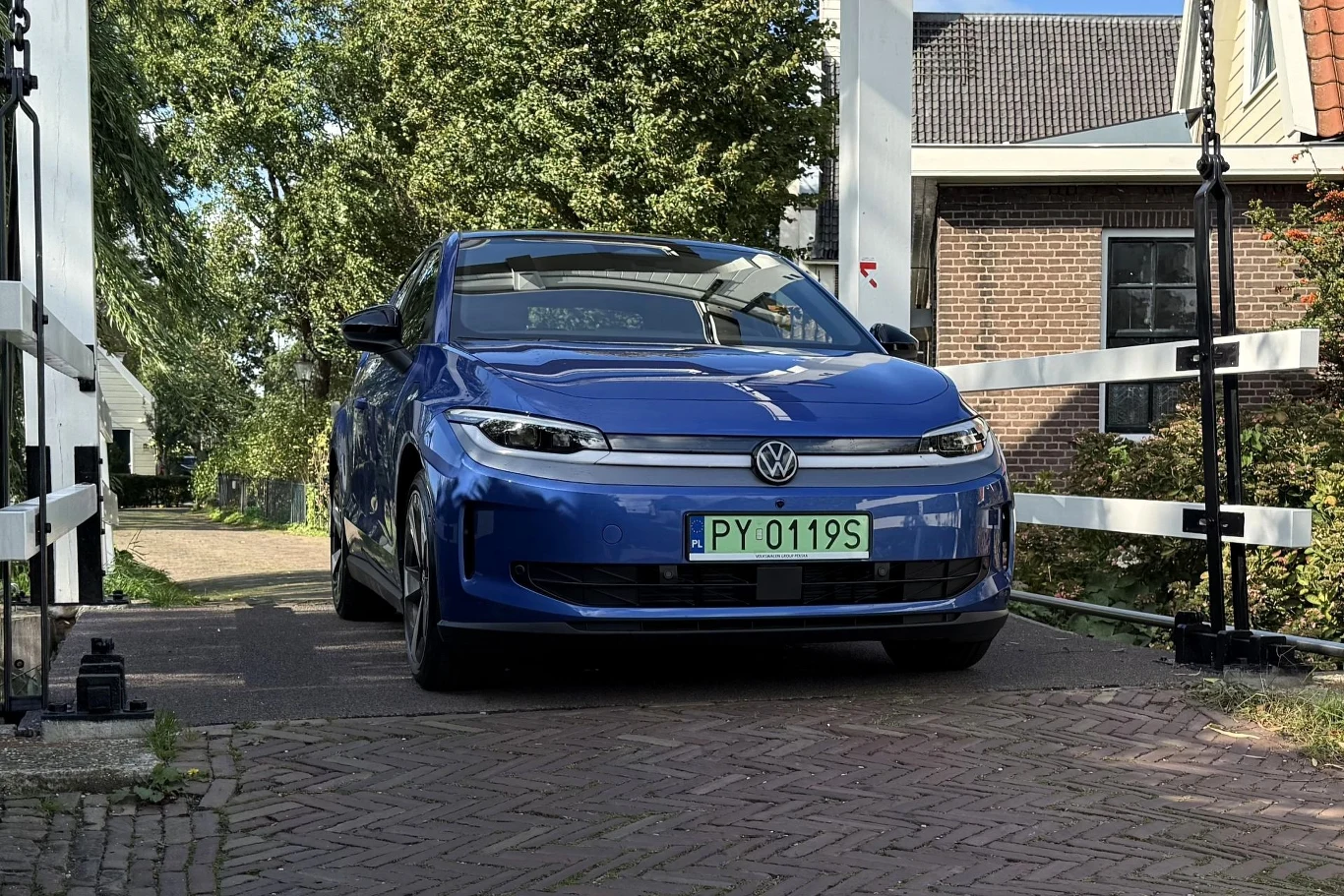 Niebieski samochód marki Volkswagen z zielonymi tablicami rejestracyjnymi stoi na brukowanym moście, otoczony zielenią i budynkami mieszkalnymi.