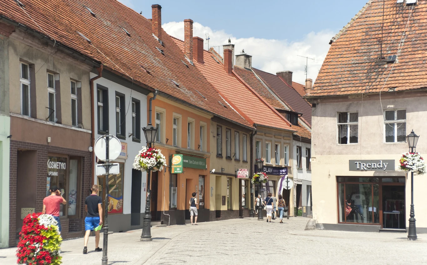 Rynek w Miliczu zachęca do spacerów Szereg kolorowych, niskich kamienic z czerwonymi dachami wzdłuż brukowanej ulicy, kilka osób spaceruje lub stoi przed witrynami sklepów, po bokach uliczne lampy ozdobione wiszącymi kwiatami.