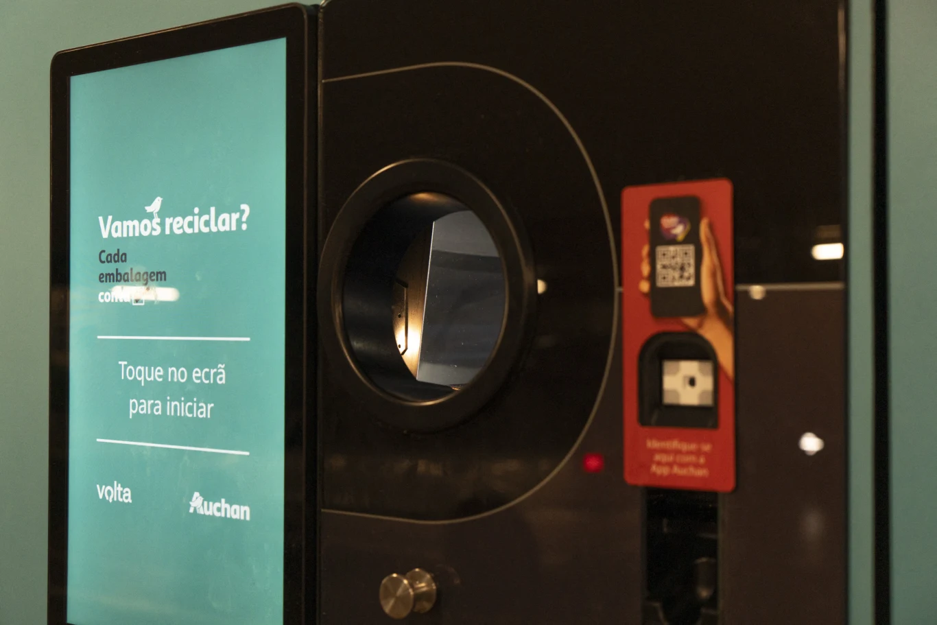 Automat do zwrotu butelek z dotykowym ekranem i otworem do przyjmowania opakowań, umieszczony przy ścianie w przestrzeni sklepowej.