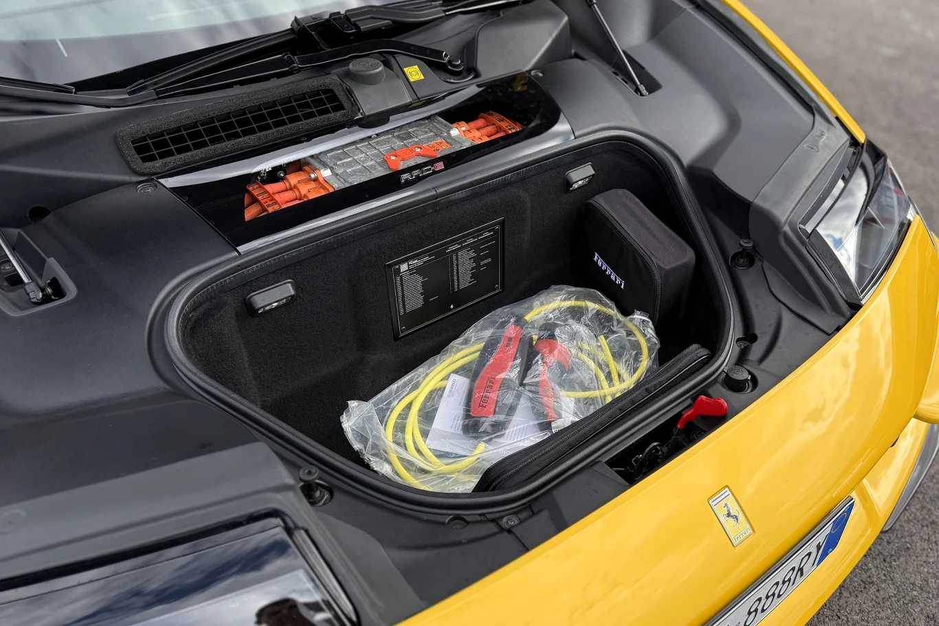 Ferrari 849 Testarossa potrai przejechać do 25 km na samym prądzie. Z przodu znajdziemy niewielki schowek na kable do ładowania i ewentualnie mały plecak lub torebkę. Nic większego się tutaj nie zmieści. Bagażnik przedni żółtego sportowego samochodu Ferrari zawierający kable rozruchowe i ładowania oraz czarną torbę serwisową.