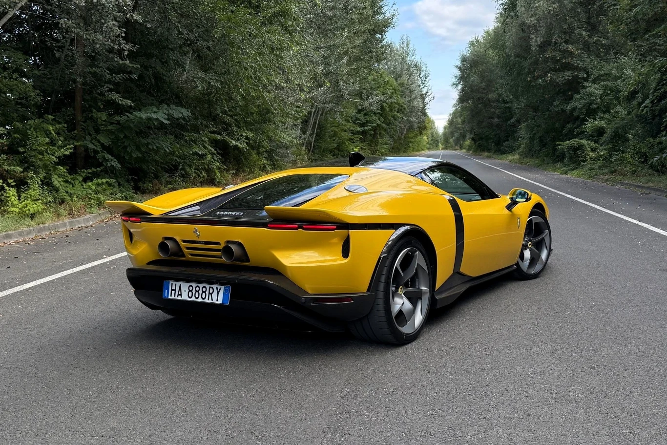 Ferrari 849 Testarossa generuje 1050 KM mocy systemowej, o 50 KM więcej niż poprzednik. Żółty sportowy samochód Ferrari SF90 Stradale stoi na pustej, asfaltowej drodze otoczonej drzewami, z wyraźnie widoczną linią maski i opływową sylwetką auta.