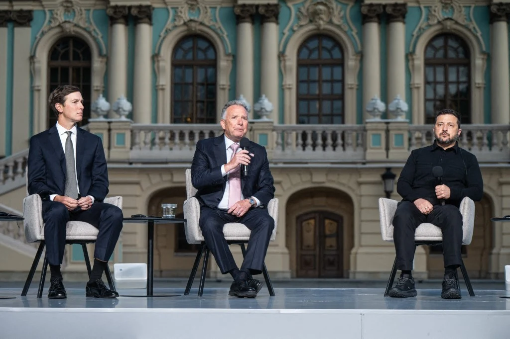 Wspólna konferencja prasowa (od prawej) Wołodymyra Zełenskiego, Steve'a Witkoffa i Jareda Kushnera Trzech mężczyzn siedzi na scenie w eleganckich fotelach, ubrani w garnitury i swetry, pośrodku jeden z nich trzyma mikrofon, w tle neobarokowa fasada budynku z dużymi oknami i kolumnami.