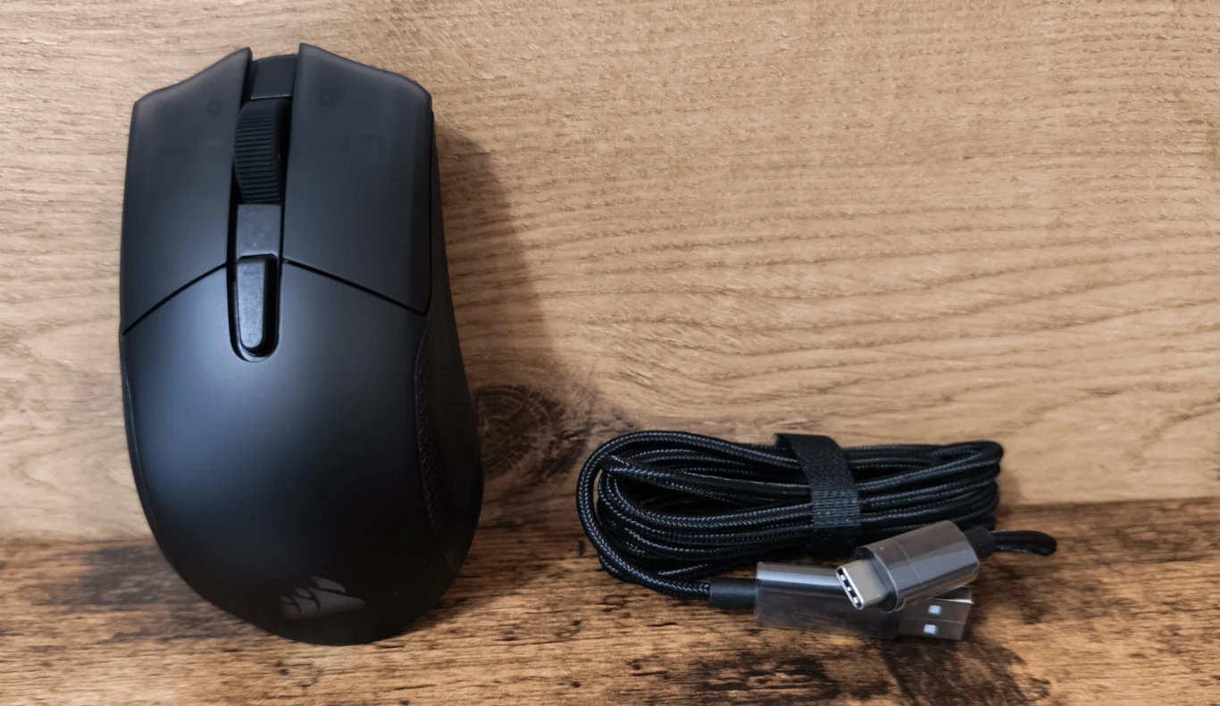 Corsair Harpoon V2 Wireless Czarna mysz komputerowa o ergonomicznym kształcie ustawiona obok zwiniętego, czarnego przewodu USB z metalową końcówką.