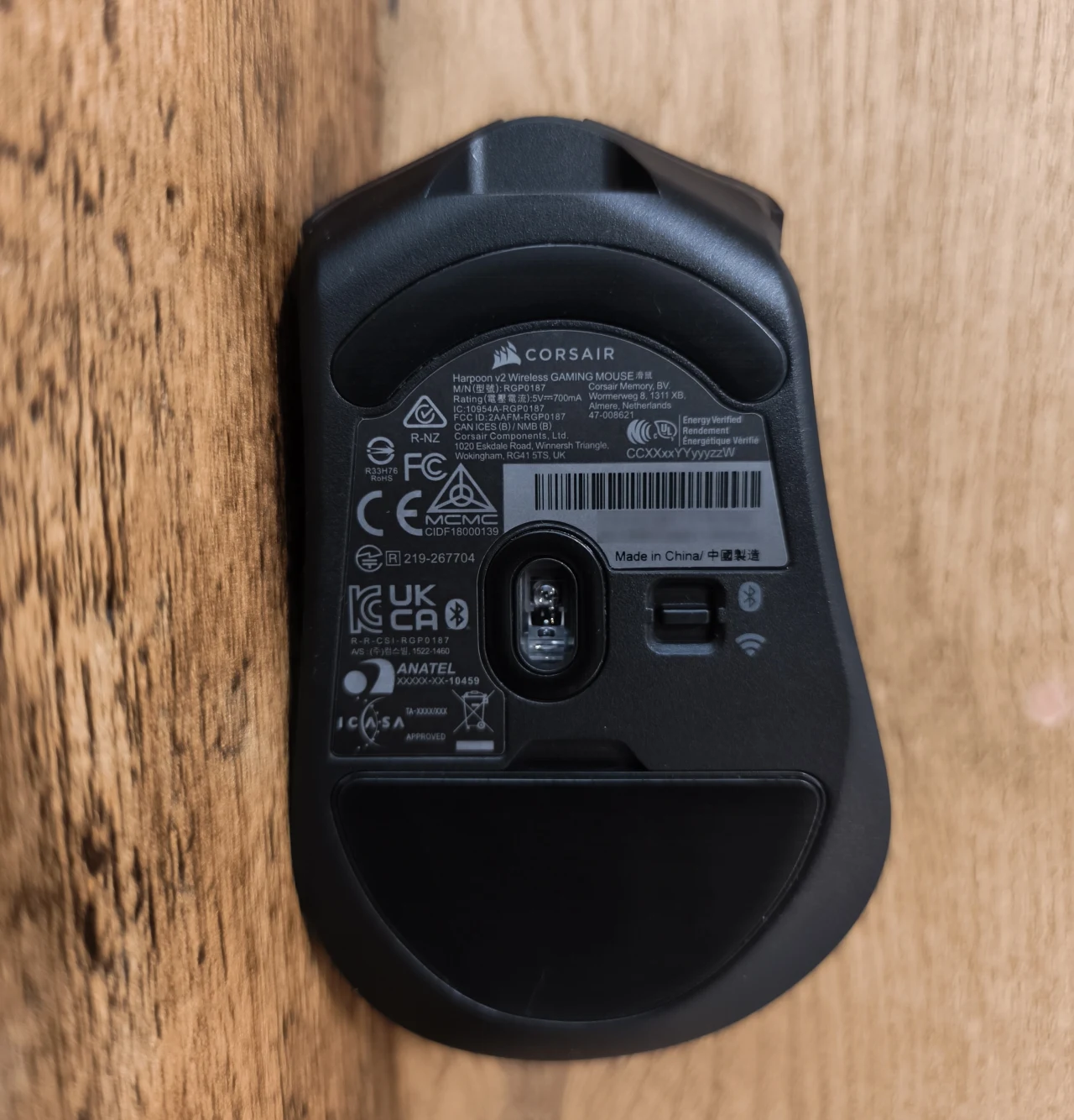 Corsair Harpoon V2 Wireless Czarna mysz komputerowa Corsair odwrócona spodem do góry, widoczne etykiety techniczne, dioda optyczna oraz przełącznik, całość na tle drewnianej powierzchni.