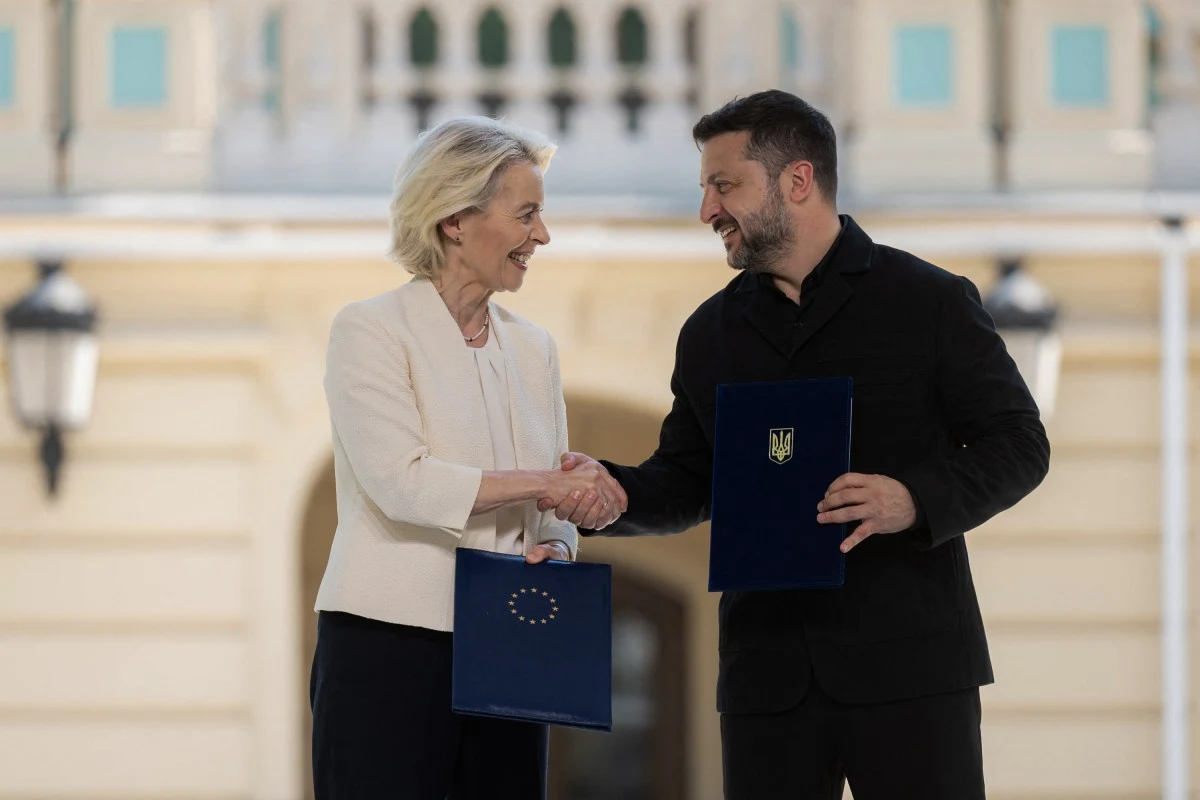 Przewodnicząca Komisji Europejskiej Ursula von der Leyen i Wołodymyr Zełenski Dwie osoby wymieniają uścisk dłoni, trzymając teczki z symbolami Unii Europejskiej oraz Ukrainy, na tle jasnego budynku.