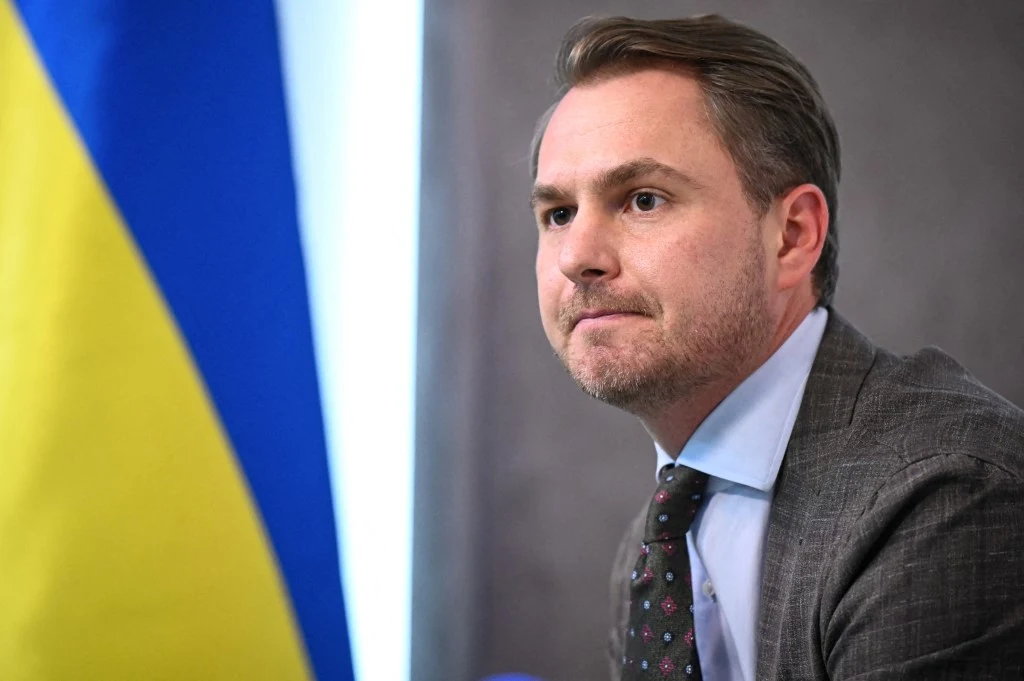 Były prokurator generalny Ukrainy Rusłan Krawczenko Mężczyzna w garniturze i krawacie siedzi przy stole, w tle fragment niebiesko-żółtej flagi.