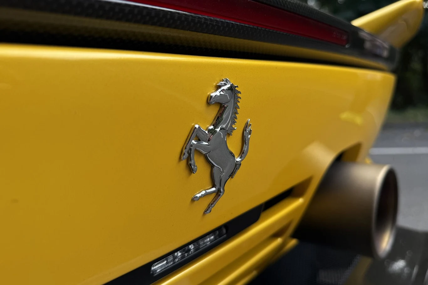 Ferrari 849 Testarossa jest wyposażone w system FIVE (Ferrari Integrated Vehicle Estimator), który tworzy cyfrowego bliźniaka naszego samochodu. Ten powtarza zachowanie prawdziwego auta w czasie rzeczywistym. Srebrny emblemat ferrari na żółtym lakierze sportowego samochodu, obok masywnej końcówki wydechu.