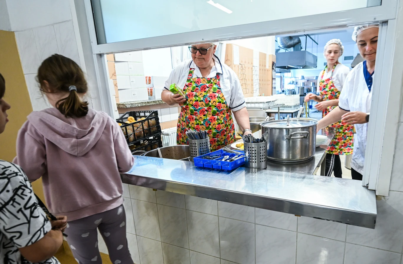 Pracownice stołówki szkolnej w fartuchach wydają obiady dzieciom stojącym przy okienku wydawczym, na blacie widoczne są garnki i naczynia.