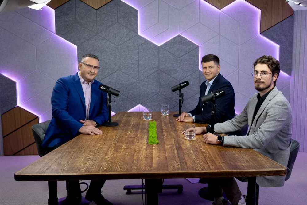 Trzech mężczyzn siedzących przy drewnianym stole w nowoczesnym studiu do nagrywania podcastów, każdy z nich ma przed sobą mikrofon i szklankę wody, w tle geometryczne panele ścienne podświetlone na fioletowo.