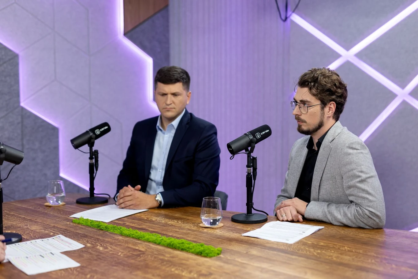 Dwóch mężczyzn w garniturach siedzi przy stole w profesjonalnym studiu nagraniowym, przed mikrofonami i kartkami z notatkami, w tle nowoczesna geometryczna ściana z podświetleniem LED.